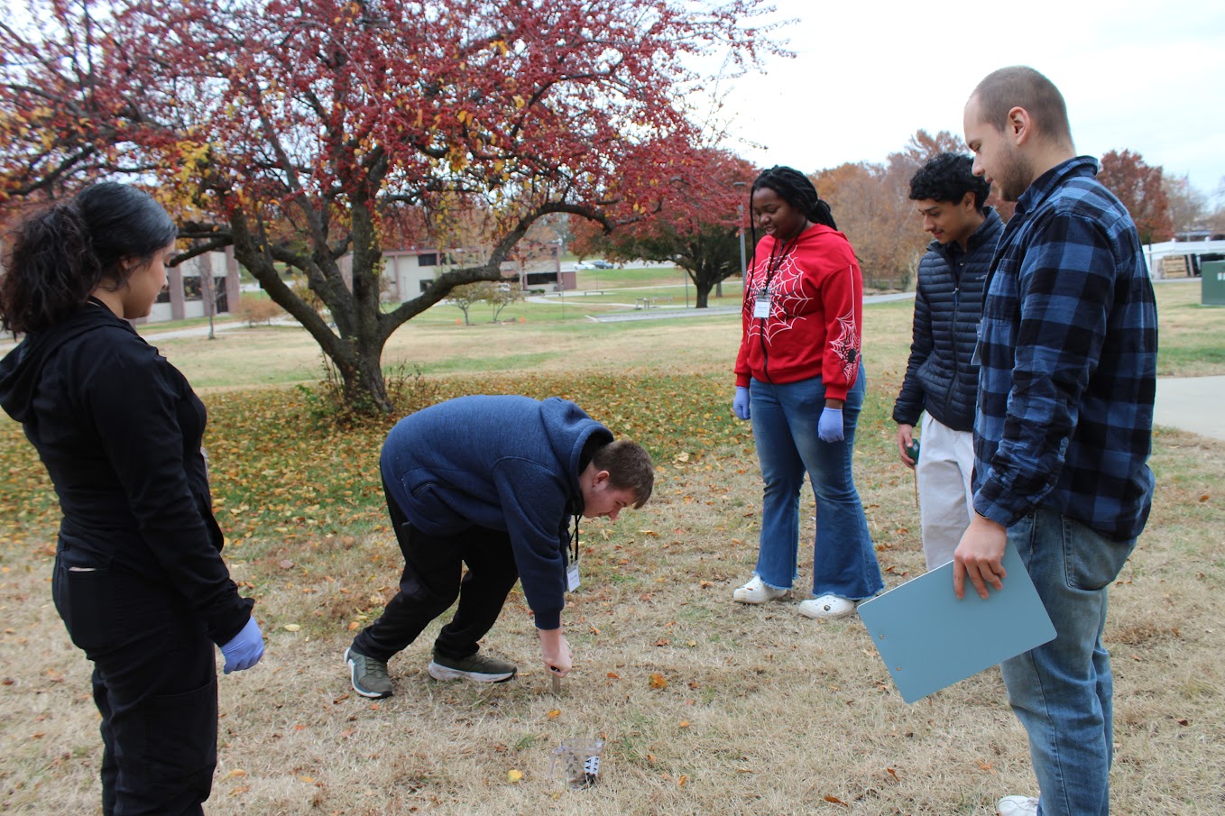 5 students standing in the grass, one leans down to take a soil sample