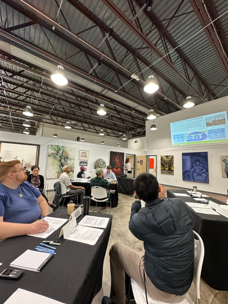 People sit at tables with a women speaking and pointing to a slide on the screen